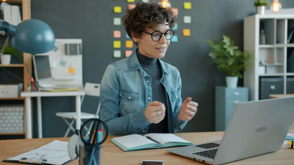 Frau mit Brille und Jeansjacke spricht gestikulierend in einen Laptop auf ihrem Schreibtisch, im Hintergrund Regale, Pflanzen und bunte Haftnotizen an der Wand